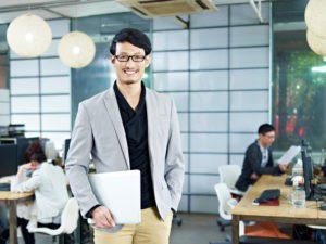 Picture of young asian entrepreneur standing in office with laptop computer under arm.