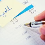 A fountain pen checks a financial or business document with an adhesive note reading "payroll" attached to it.