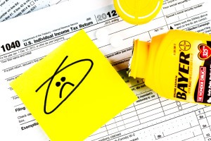 Form 1040 pictured with a bottle of Bayer aspirin and a doodle of a frowning face on a sticky note.